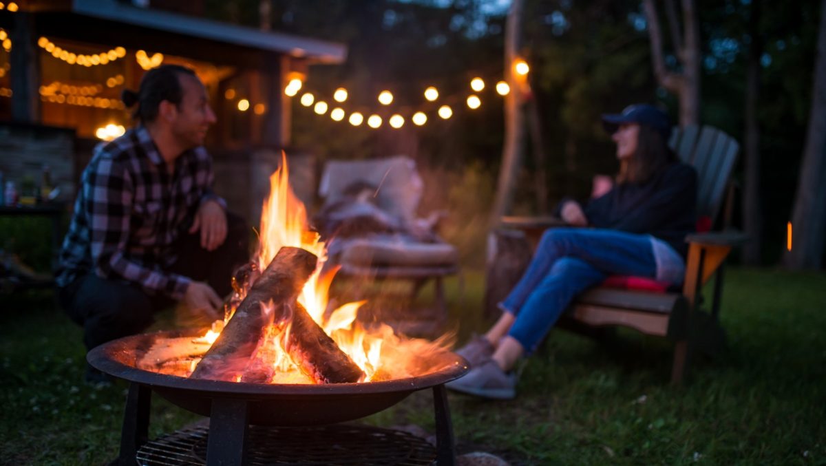 A man, woman, and child sitting around a burning fire pit in the woods with fairy lights blurred in the background