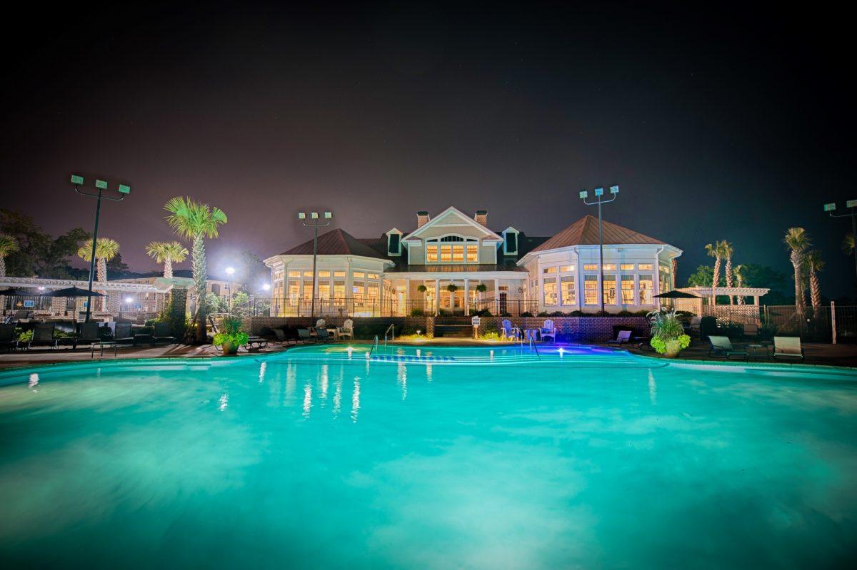 Resort-style pool with lounge chairs around the pool deck and the clubhouse in the background at night time.